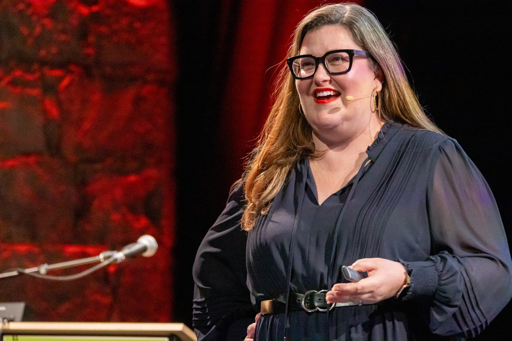 Avery Swartz wears black rimmed glasses, a navy dress, and smiles while on stage speaking